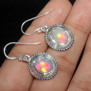 May include: A pair of silver-coloured drop earrings. Each earring has an oval, iridescent opal stone set in a decorative, textured frame. The opals show a mix of pink, yellow, and green hues. The earrings are displayed on a hand.