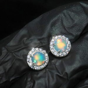 May include: A pair of silver earrings featuring a round opal gemstone set in a halo of smaller clear gemstones.