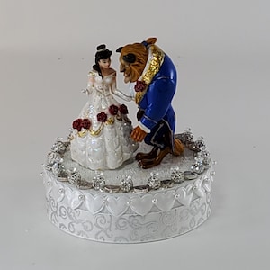 May include: A decorative figurine featuring Belle and the Beast from Beauty and the Beast. Belle wears a white dress with red roses, and the Beast is in a blue suit. The base is white with silver accents and heart patterns.