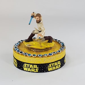 May include: A Star Wars themed figurine of Obi-Wan Kenobi in a fighting pose, holding a blue lightsaber. The figure is on a yellow base with blue gems and a black ribbon with "Star Wars" text.