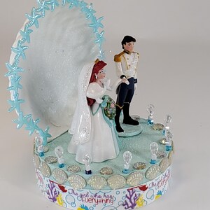 K&ouml;nnte beinhalten: Eine Tortenaufsatz f&uuml;r eine Hochzeit mit Braut- und Br&auml;utigamfiguren. Die Braut tr&auml;gt ein wei&szlig;es Kleid und einen Schleier und h&auml;lt einen Blumenstrau&szlig;. Der Br&auml;utigam tr&auml;gt eine wei&szlig;e und goldene Uniform. Die Basis ist mit Muscheln und dem Text "The girl who has everything" verziert.