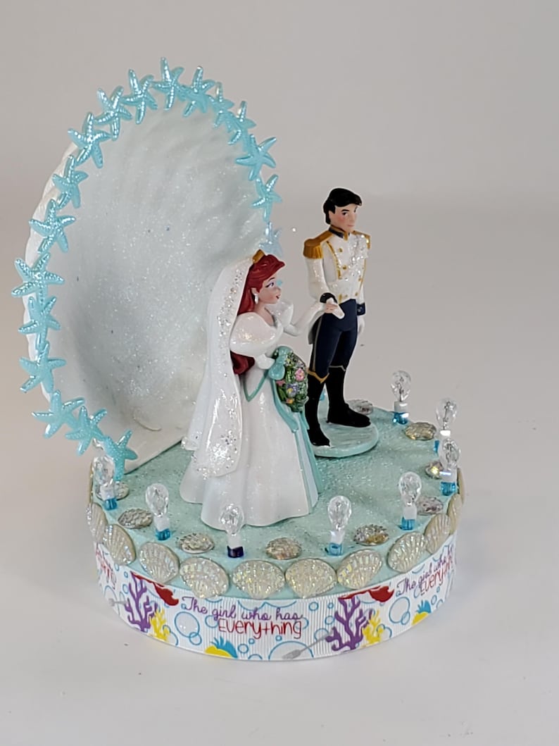 K&ouml;nnte beinhalten: Eine Tortenaufsatz f&uuml;r eine Hochzeit mit Braut- und Br&auml;utigamfiguren. Die Braut tr&auml;gt ein wei&szlig;es Kleid und einen Schleier und h&auml;lt einen Blumenstrau&szlig;. Der Br&auml;utigam tr&auml;gt eine wei&szlig;e und goldene Uniform. Die Basis ist mit Muscheln und dem Text "The girl who has everything" verziert.