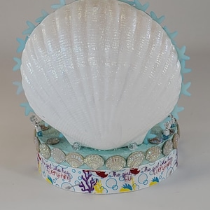 K&ouml;nnte beinhalten: Eine dekorative Muschel mit einer wei&szlig;en, schillernden Oberfl&auml;che, verziert mit hellblauen Seesternen. Der Sockel hat ein Band mit dem Text "The girl who has everything" und Muschelschmuck. Das Design hat ein maritimes Thema.
