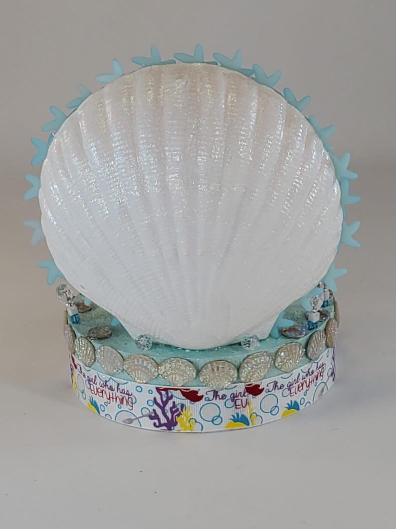 K&ouml;nnte beinhalten: Eine dekorative Muschel mit einer wei&szlig;en, schillernden Oberfl&auml;che, verziert mit hellblauen Seesternen. Der Sockel hat ein Band mit dem Text "The girl who has everything" und Muschelschmuck. Das Design hat ein maritimes Thema.