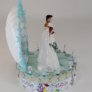 K&ouml;nnte beinhalten: Dekorative Figur mit einer Muschel mit blauen Akzenten und einem Paar in Hochzeitskleidung. Der Sockel ist mit Muscheln und dem Text "The girl who has everything" verziert.