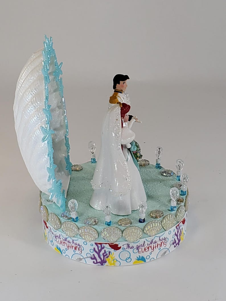 K&ouml;nnte beinhalten: Dekorative Figur mit einer Muschel mit blauen Akzenten und einem Paar in Hochzeitskleidung. Der Sockel ist mit Muscheln und dem Text "The girl who has everything" verziert.