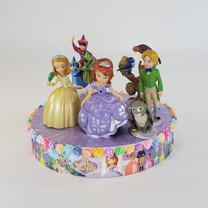 May include: A cake topper featuring characters from the animated series "Sofia the First." The topper includes figurines of Sofia, other characters, and a rabbit, arranged on a purple base with a ribbon that says "Sofia the First."