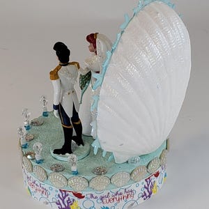K&ouml;nnte beinhalten: Eine Tortenaufsatz f&uuml;r eine Hochzeit mit Braut- und Br&auml;utigamfiguren, einer gro&szlig;en wei&szlig;en Muschel und dem Text "The girl who has everything". Die Basis ist mit Muscheln und Kristallen verziert.