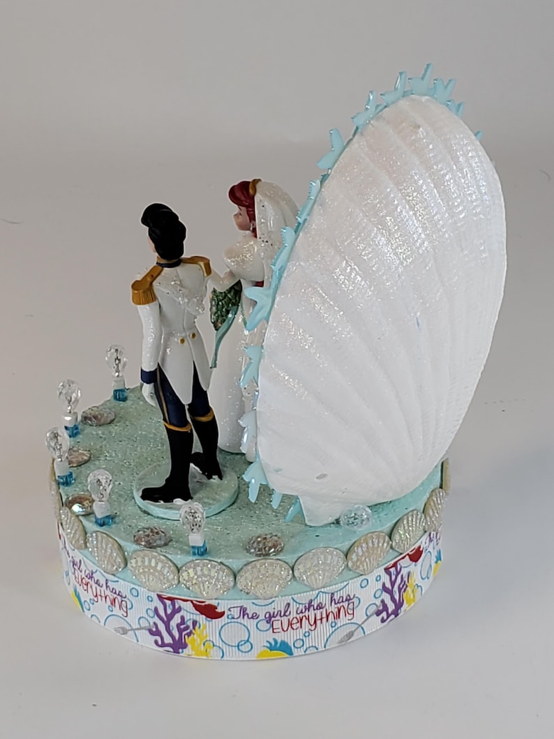 K&ouml;nnte beinhalten: Eine Tortenaufsatz f&uuml;r eine Hochzeit mit Braut- und Br&auml;utigamfiguren, einer gro&szlig;en wei&szlig;en Muschel und dem Text "The girl who has everything". Die Basis ist mit Muscheln und Kristallen verziert.