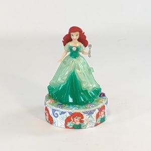 May include: A green and light green dress with a sparkly bodice and a red haired Disney princess figurine holding a silver fork. The figurine is on a round base with a colorful design of the princess.