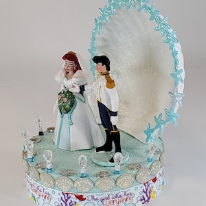 K&ouml;nnte beinhalten: Eine Tortenaufsatz f&uuml;r eine Hochzeit mit Braut- und Br&auml;utigamfiguren. Die Braut tr&auml;gt ein wei&szlig;es Kleid und einen Schleier und h&auml;lt einen Blumenstrau&szlig;. Der Br&auml;utigam tr&auml;gt eine wei&szlig;e und goldene Uniform. Die Basis ist mit Muscheln und dem Text "The girl who has Everything" verziert.