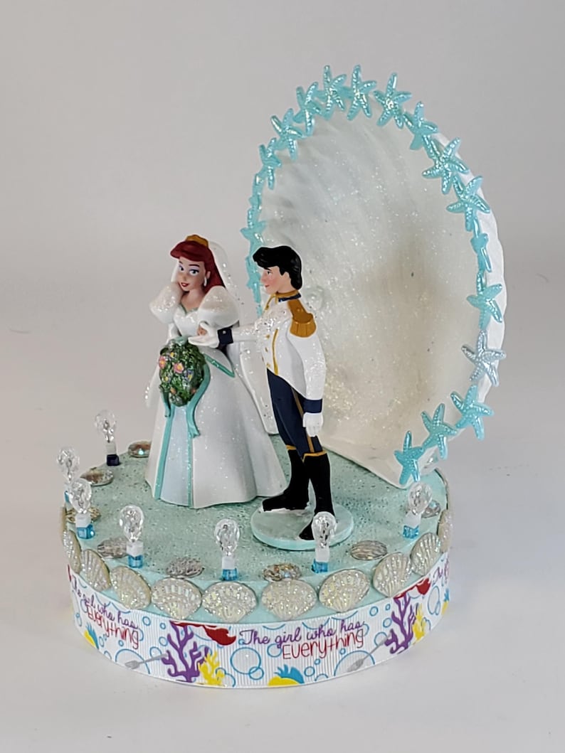 K&ouml;nnte beinhalten: Eine Tortenaufsatz f&uuml;r eine Hochzeit mit Braut- und Br&auml;utigamfiguren. Die Braut tr&auml;gt ein wei&szlig;es Kleid und einen Schleier und h&auml;lt einen Blumenstrau&szlig;. Der Br&auml;utigam tr&auml;gt eine wei&szlig;e und goldene Uniform. Die Basis ist mit Muscheln und dem Text "The girl who has Everything" verziert.