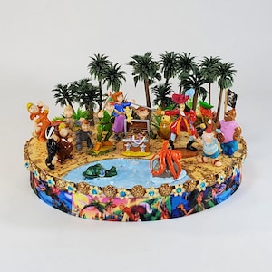 May include: A decorative Peter Pan-themed figurine featuring characters like Peter Pan, Captain Hook, and Tinkerbell. The scene includes palm trees, a pond, and a treasure chest, all set on a detailed base with a colorful ribbon border.