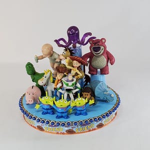 May include: A blue, tiered cake decorated with various Toy Story character figurines. The cake features a blue base with a gold beaded border and the words "You've Got a Friend in Me."