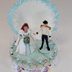 K&ouml;nnte beinhalten: Eine Tortenaufsatz f&uuml;r eine Hochzeit mit Braut- und Br&auml;utigamfiguren. Die Braut tr&auml;gt ein wei&szlig;es Kleid und einen Schleier und h&auml;lt einen Blumenstrau&szlig;. Der Br&auml;utigam tr&auml;gt eine wei&szlig;e Jacke und eine schwarze Hose. Die Basis ist mit Muscheln und dem Text "The girl who has Everything" verziert.