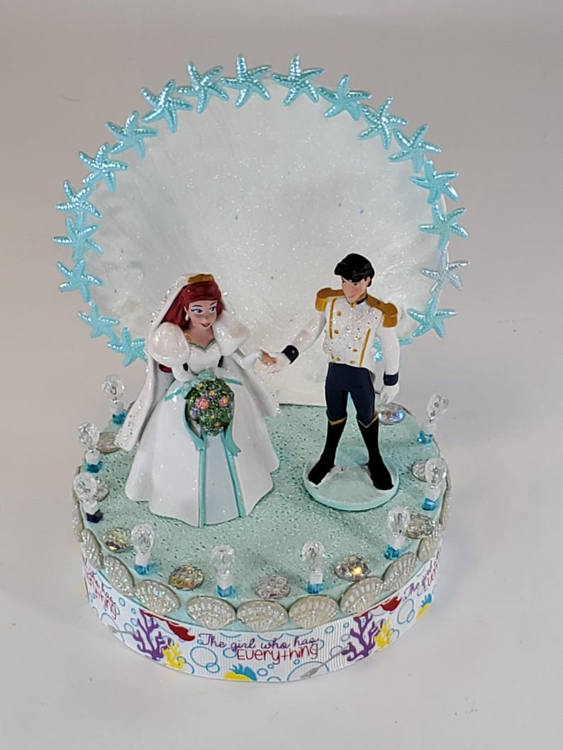 K&ouml;nnte beinhalten: Eine Tortenaufsatz f&uuml;r eine Hochzeit mit Braut- und Br&auml;utigamfiguren. Die Braut tr&auml;gt ein wei&szlig;es Kleid und einen Schleier und h&auml;lt einen Blumenstrau&szlig;. Der Br&auml;utigam tr&auml;gt eine wei&szlig;e Jacke und eine schwarze Hose. Die Basis ist mit Muscheln und dem Text "The girl who has Everything" verziert.