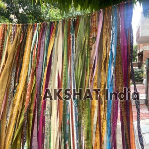 May include: A colorful fabric garland with strips of various colors and patterns, including gold, green, blue, and pink. The garland is hanging from a tree branch and has the text "AKSHAT India" printed on it.