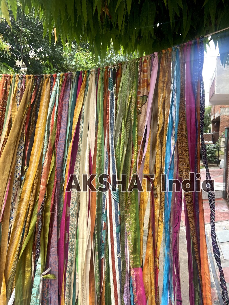 May include: A colorful fabric garland with strips of various colors and patterns, including gold, green, blue, and pink. The garland is hanging from a tree branch and has the text "AKSHAT India" printed on it.