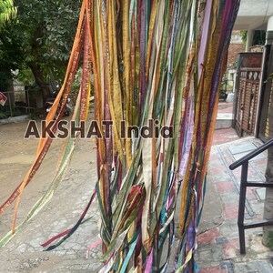 May include: A colorful curtain made of many strips of fabric, hanging from a string. The fabric is in various shades of yellow, orange, red, green, blue, purple, and brown. The curtain is hanging in front of a brick wall.