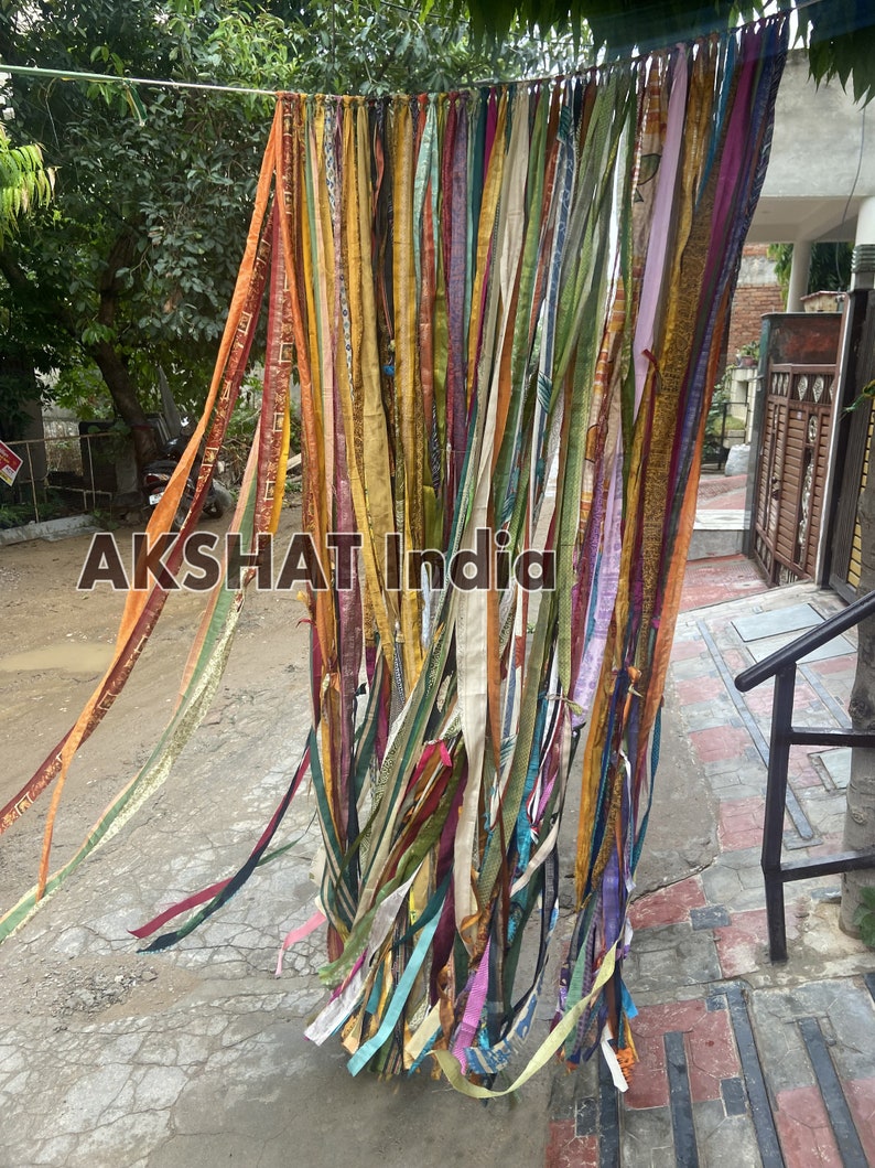 May include: A colorful curtain made of many strips of fabric, hanging from a string. The fabric is in various shades of yellow, orange, red, green, blue, purple, and brown. The curtain is hanging in front of a brick wall.