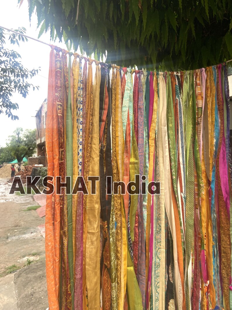 May include: A colorful fabric curtain made of many strips of fabric in various colors and patterns, including orange, yellow, green, blue, and purple. The fabric is tied together at the top and hangs down in a cascade of color. The text "AKSHAT India" is visible at the bottom of the image.
