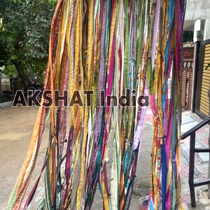 May include: A colorful curtain made of strips of fabric in various colors, including yellow, orange, pink, purple, blue, and green. The fabric strips are hanging from a string and are arranged in a cascading pattern. The text "AKSHAT India" is visible in the center of the image.