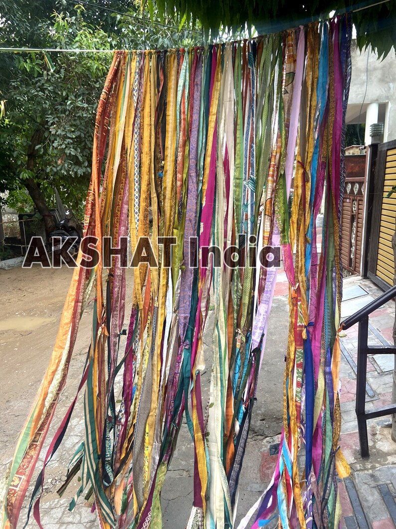 May include: A colorful curtain made of strips of fabric in various colors, including yellow, orange, pink, purple, blue, and green. The fabric strips are hanging from a string and are arranged in a cascading pattern. The text "AKSHAT India" is visible in the center of the image.