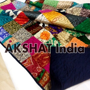 Może przedstawiać: Kolorowa kołdra patchworkowa z granatowym tyłem. Kołdra wykonana jest z wielu różnych tkanin i wzorów, w tym kwiatowych, geometrycznych i paisley. Kołdra jest złożona na pół, a na niej widoczny jest napis "AKSHAT India".