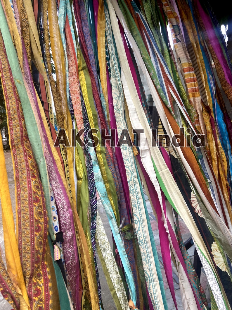 May include: A wall decorated with colorful fabric strips hanging from the top. The strips are of various lengths and widths, and they are arranged in a random pattern. The fabric is a mix of different colors, patterns, and textures. The text "AKSHAT India" is visible in the center of the image.