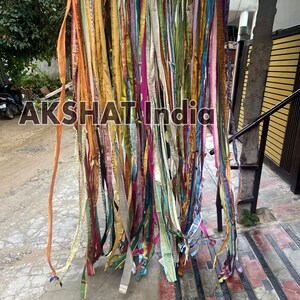 May include: A colorful curtain made of many strips of fabric hanging from a string. The fabric is in various colors, including orange, yellow, pink, blue, green, and purple. The curtain is hanging in front of a brick wall.
