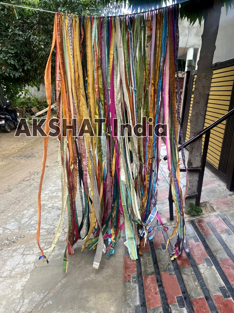 May include: A colorful curtain made of many strips of fabric hanging from a string. The fabric is in various colors, including orange, yellow, pink, blue, green, and purple. The curtain is hanging in front of a brick wall.