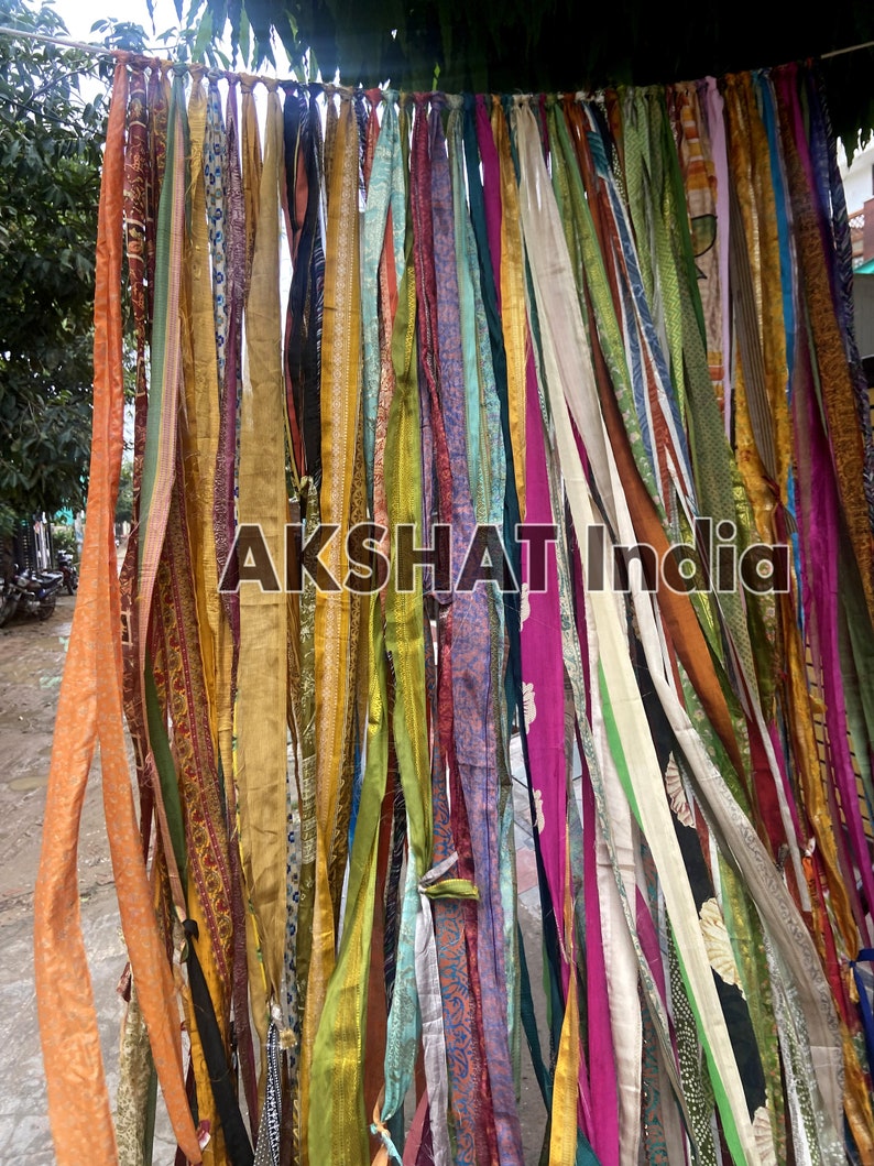 May include: A colorful curtain made of many strips of fabric in various colors, including orange, yellow, green, blue, purple, and pink. The fabric strips are tied together at the top and hang down in a cascade. The text "AKSHAT India" is visible in the center of the image.