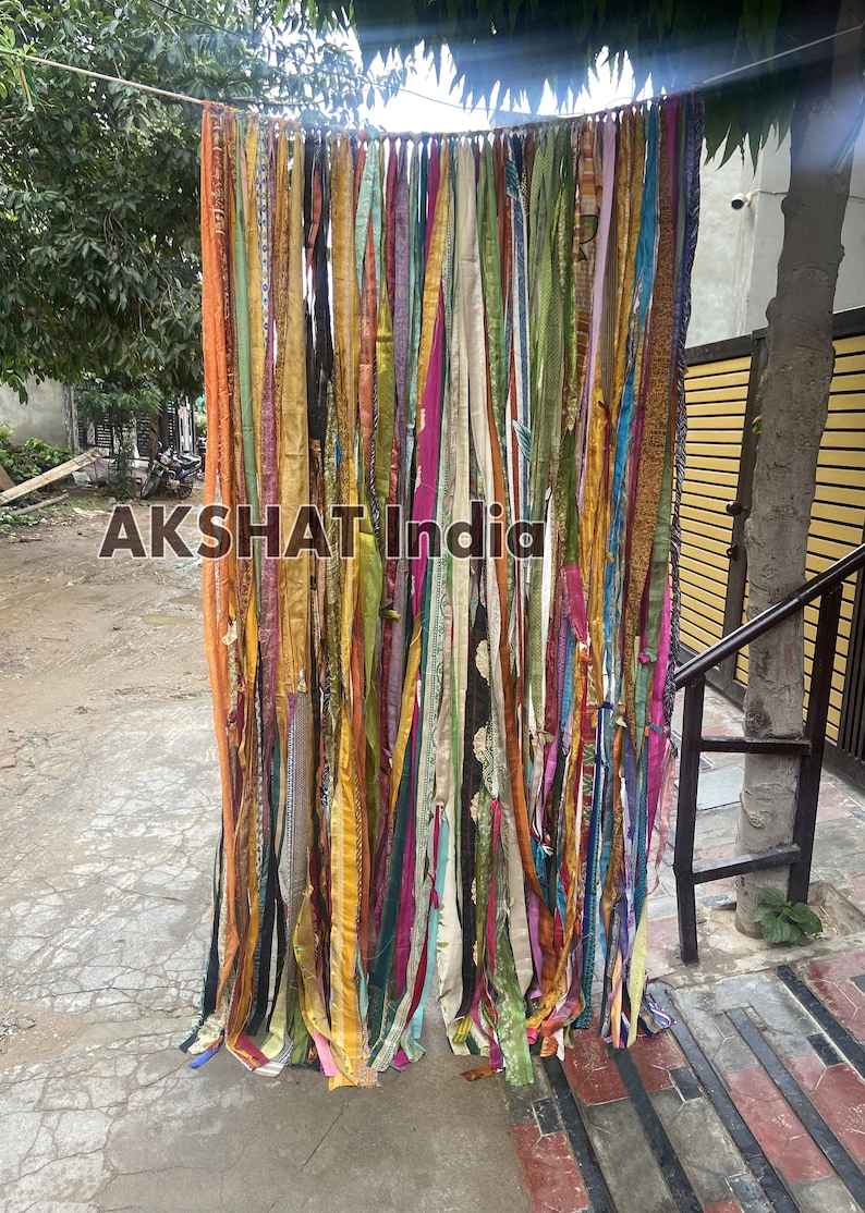 May include: A colorful curtain made of many strips of fabric, hanging from a string. The fabric is in various shades of green, yellow, orange, pink, purple, and blue. The curtain is hanging in front of a yellow wall and a tree.