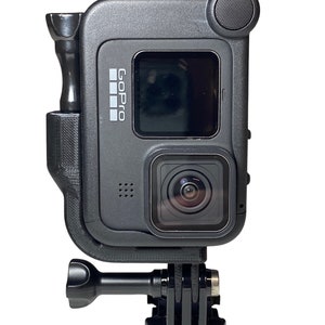 May include: Black GoPro action camera with a black protective case and a GoPro logo on the side. The camera has a small screen on the back and a lens on the front. The camera is attached to a black GoPro mount.