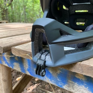 Puede incluir: Primer plano de un casco de bicicleta negro con un soporte de c&aacute;mara integrado. El casco tiene un acabado mate y un dise&ntilde;o texturizado. El soporte de c&aacute;mara est&aacute; ubicado en el lateral del casco y est&aacute; hecho de pl&aacute;stico negro.