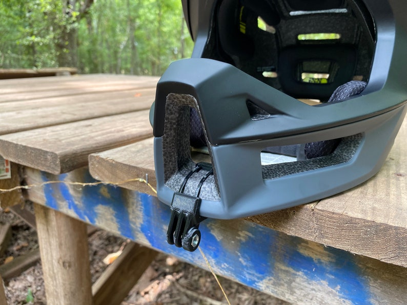 Puede incluir: Primer plano de un casco de bicicleta negro con un soporte de c&aacute;mara integrado. El casco tiene un acabado mate y un dise&ntilde;o texturizado. El soporte de c&aacute;mara est&aacute; ubicado en el lateral del casco y est&aacute; hecho de pl&aacute;stico negro.