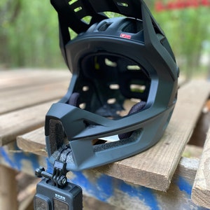 Puede incluir: Un casco integral negro con visera y una c&aacute;mara GoPro fijada en el lateral. El casco est&aacute; sobre una superficie de madera.