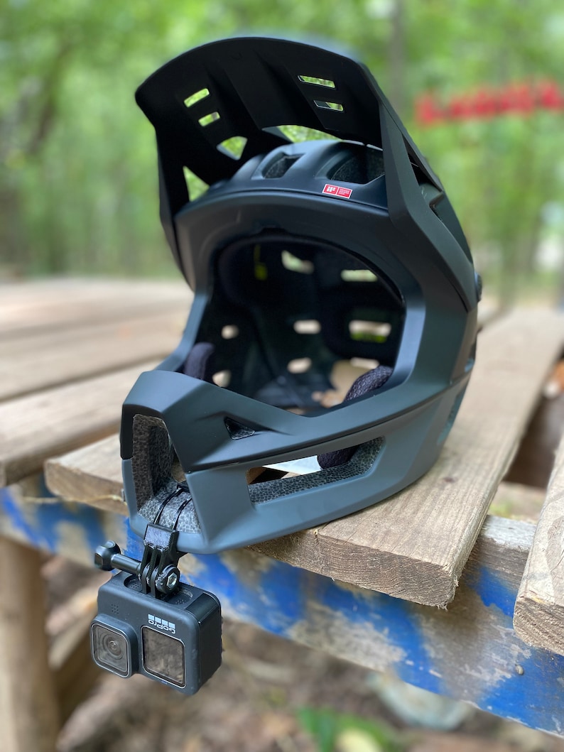 Puede incluir: Un casco integral negro con visera y una c&aacute;mara GoPro fijada en el lateral. El casco est&aacute; sobre una superficie de madera.