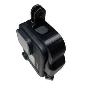 May include: Black GoPro camera with a black protective case and a black mount on top. The camera has a small screen on the back and a lens on the front.