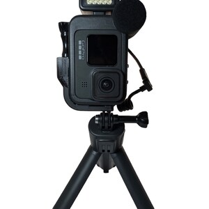 May include: A black GoPro camera mounted on a small black tripod with a black microphone and a small black LED light attached to the top of the camera.