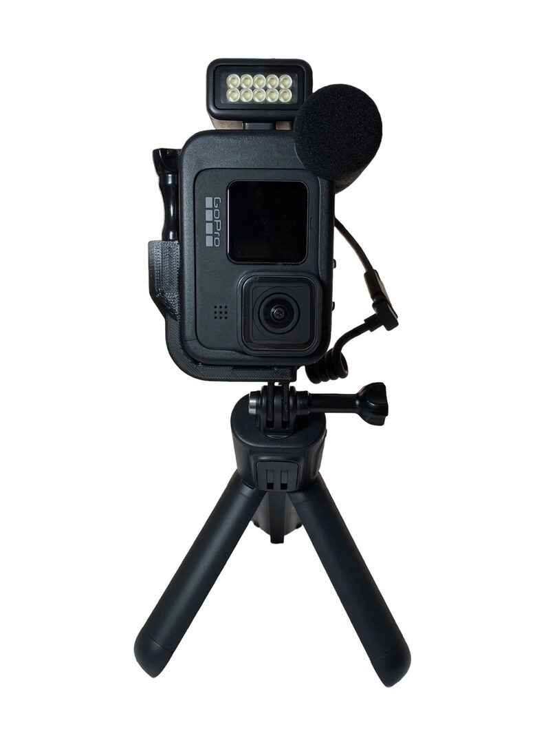 May include: A black GoPro camera mounted on a small black tripod with a black microphone and a small black LED light attached to the top of the camera.