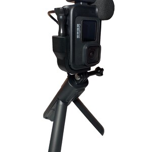 May include: A black GoPro camera mounted on a small black tripod with a black microphone and a black LED light attached to the top.