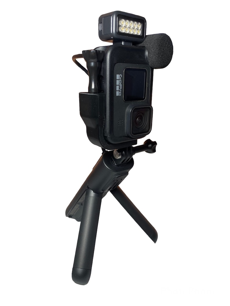 May include: A black GoPro camera mounted on a small black tripod with a black microphone and a black LED light attached to the top.
