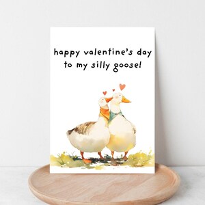 May include: A Valentine's Day card featuring a watercolor illustration of two geese with hearts above their heads. The card reads "happy valentine's day to my silly goose!" on a white background. The card is displayed on a wooden tray.