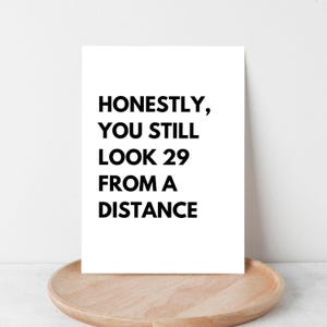 Könnte beinhalten: Eine weiße rechteckige Karte mit dem Text "HONESTLY, YOU STILL LOOK 29 FROM A DISTANCE" in Schwarz. Die Karte wird auf einem runden, hellbraunen Holztablett präsentiert.