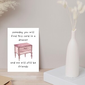 Peut inclure: Une carte blanche avec le texte "someday you will find this card in a drawer and we will still be friends." La carte pr&eacute;sente une illustration &agrave; l'aquarelle d'une table de chevet rose. Un vase blanc avec des fleurs s&eacute;ch&eacute;es est &agrave; droite.