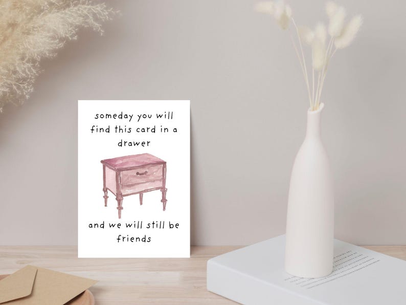 Peut inclure: Une carte blanche avec le texte "someday you will find this card in a drawer and we will still be friends." La carte pr&eacute;sente une illustration &agrave; l'aquarelle d'une table de chevet rose. Un vase blanc avec des fleurs s&eacute;ch&eacute;es est &agrave; droite.
