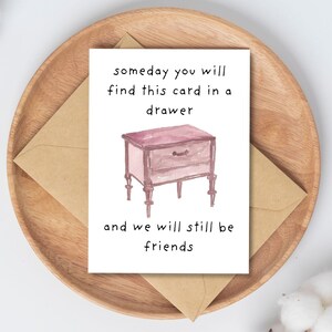 Peut inclure: Carte de v&oelig;ux avec une illustration &agrave; l'aquarelle d'une table de chevet et le texte "someday you will find this card in a drawer and we will still be friends." La carte est sur une assiette en bois avec une enveloppe brune.