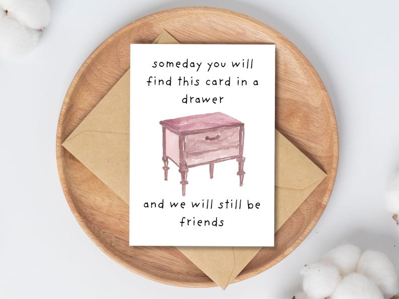 Peut inclure: Carte de v&oelig;ux avec une illustration &agrave; l'aquarelle d'une table de chevet et le texte "someday you will find this card in a drawer and we will still be friends." La carte est sur une assiette en bois avec une enveloppe brune.