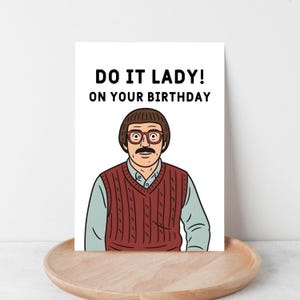 May include: A greeting card with the text "DO IT LADY! ON YOUR BIRTHDAY" above a cartoon illustration of a man wearing glasses, a red sweater vest, and a light blue shirt. The card is on a wooden tray.